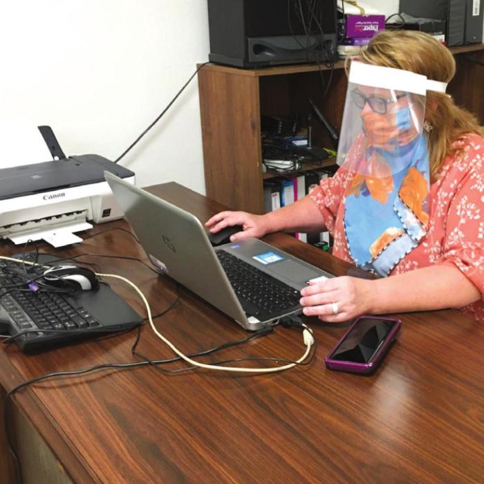 Carol Ann Thacker, outfitted with her shield and neck mask, sits at her desk last Monday. NVCC had four desks donated to them from a Houston office which closed. The desks had been sitting in a barn because there was no room for them. NVCC appreciates Chip Bubela and Coy Romine who picked up the desks and moved them in.