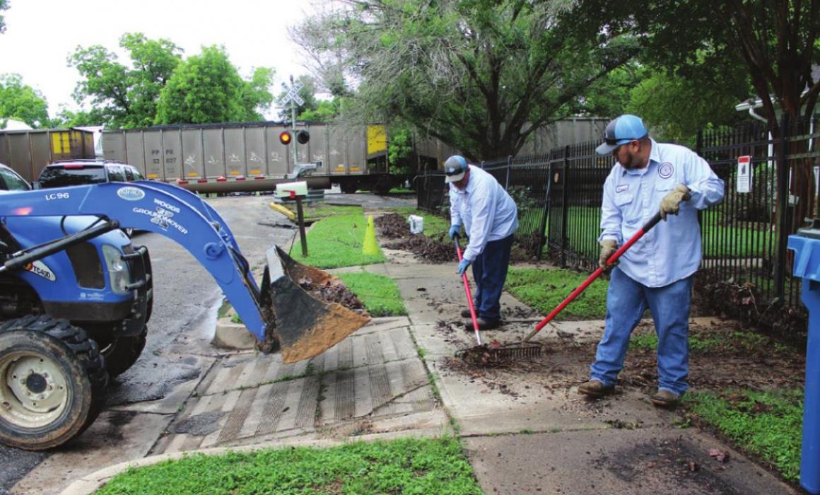 City employees Derrick Esquivel, right, and Tim Jackson, center, work to clean up storm debris that was piled up on the sidewalk of Monroe St. in La Grange. The fence at that home was damaged in several areas as well. In the background, a train goes by, near where a portion of the track had to be repaired overnight after heavy rains flooded the area. On the tractor was Don Jakobeit. Photo by Jeff Wick