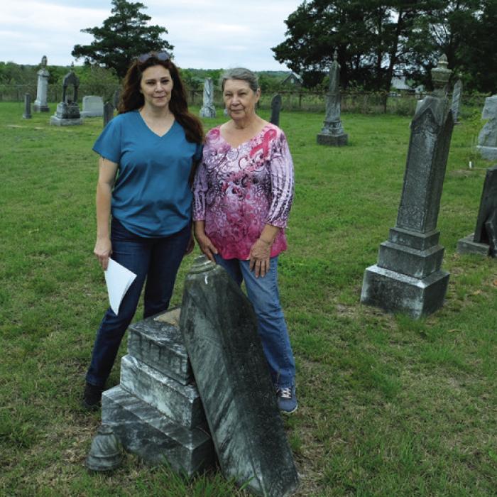 Shan Jakobeit, left, and her mother Connie stand by the grave of Caroline Otto, who died in 1898. The top of the gravestone has broken away from the leaning pedestal. Photo by Andy Behlen