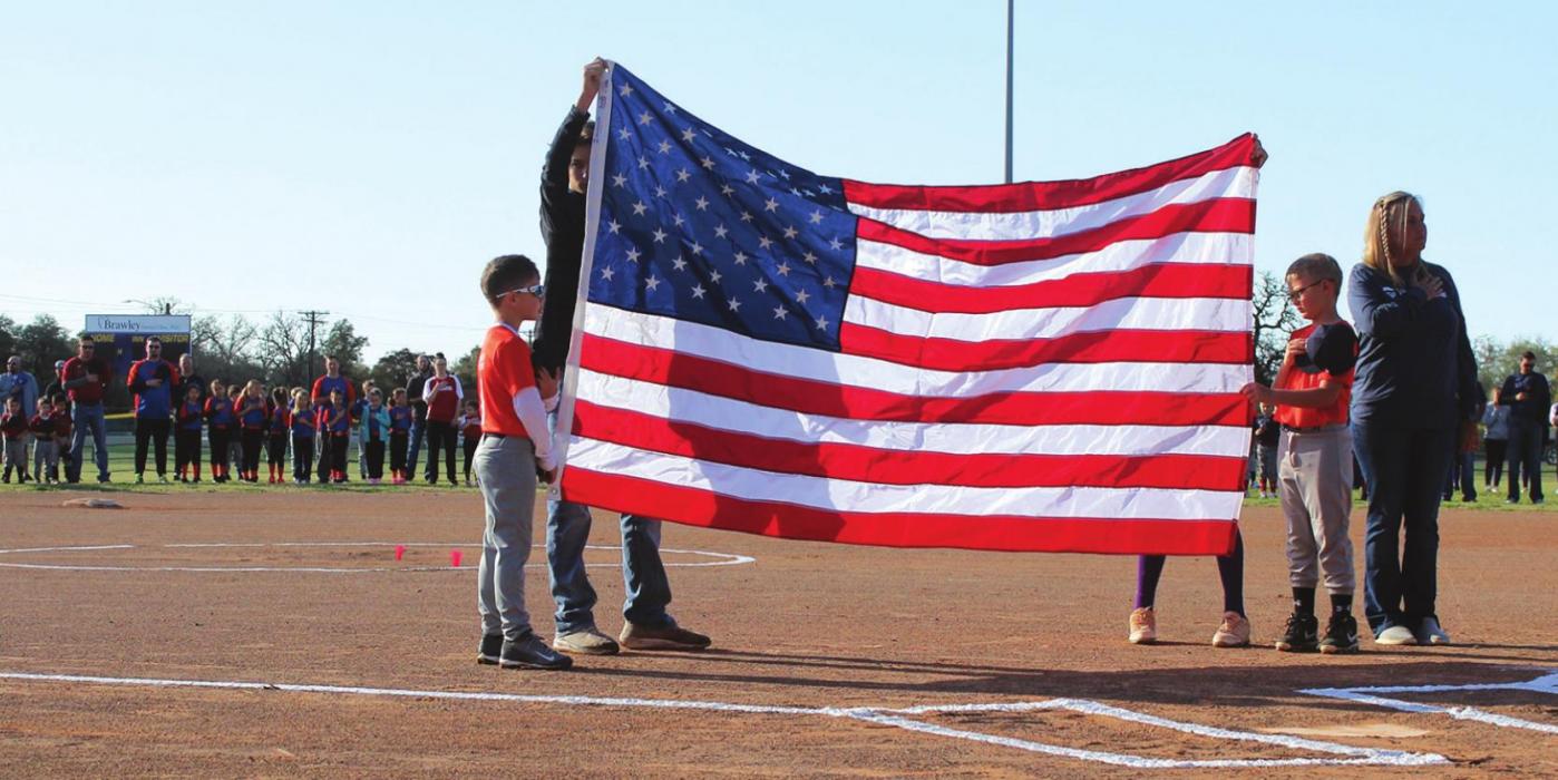 Little Leaguers hold up the flag (with a little help) during the Opening Day ceremony Saturday at the fairgrounds softball fields. Woodmen of the World donated the flag. Behind them stands some of the teams that were announced during the ceremony. Photos by Jeff Wick