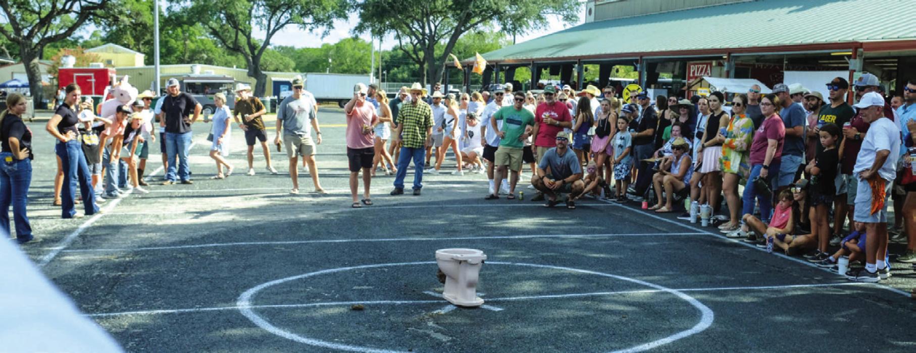 A crowd gathers around to watch the cow chip pitching contest at the Schulenburg Festival on Saturday, Aug. 5. The contest, sponsored by the Schulenburg Young Farmers, involves pitching a lump of cow manure into a toilet. Photo by Andy Behlen