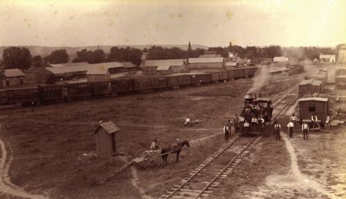This Circa 1895 photograph of the M. K. &amp; T rail yard on the eastern end of La Grange (vicinity of 600 Block East La Fayette Street) looking south with the bluff off in the distance. Louis Melcher captured this view from the roof of the M. K. T. freight depot. Train cars, warehouses, elevated tanks and open sheds like those used by lumberyards are visible (present day Farmer’s Lumber Company).